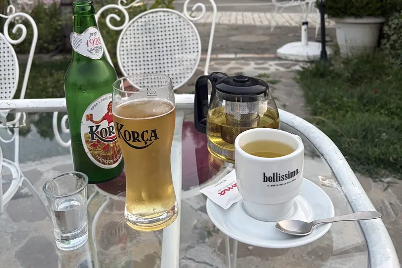 A Korça beer, a pot of tea and a cup on a glass table in the courtyard